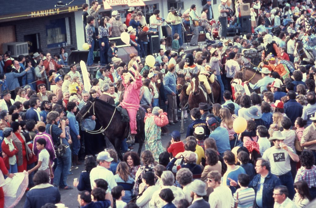 "Parade at Cajun Mardi Gras festival" 1989. Shows revelers, some riding horses, in traditional Cajun Mardi Gras outfits passing through crowds in front of Manuel's Bar, 6th Street, Mamou, Louisiana. A Cajun band plays at top.